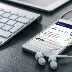 Smartfon z aplikacją KOLEO pokazuje plan podróży i opcje płatności, idealny do wyszukiwania zniżki PKP. Słuchawki i klawiatura sugerują planowanie podróży.