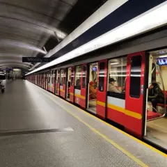 Stacja metra Młociny, do której jeździ metro w Warszawie. Czerwony pociąg z żółtym pasem stoi na peronie, czekając na pasażerów.