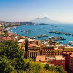 Widok na Zatokę Neapolitańską z Wezuwiuszem w tle. Miasto rozciąga się wzdłuż wybrzeża, a port pełen jest łodzi. Roma Napoli to marzenie wielu podróżników.