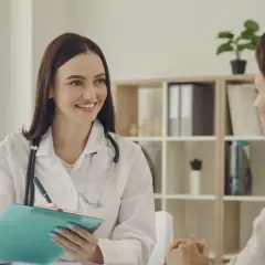 Lekarka w białym kitlu z notatnikiem i stetoskopem uśmiecha się do pacjentki. S1 Norwegia.
