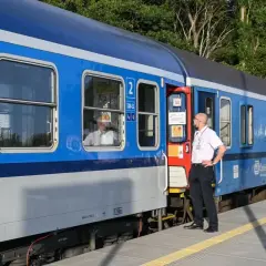 Pracownik kolei stoi przy drzwiach niebieskiego pociągu. Pasażerowie wyglądają przez okna. Czy można kupić bilet w pociągu?