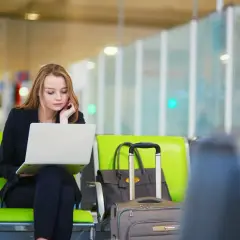 Kobieta w czarnym garniturze siedzi na zielonym krześle w poczekalni, pracując na laptopie. Obok niej walizka i torba. Może to być odwołanie rezerwacji.