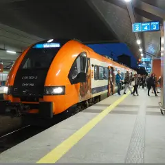 Pomarańczowy pociąg do krakowskiego lotniska na peronie. Ludzie wsiadają i wysiadają.
