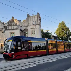 Nowoczesny tramwaj pabianice z czerwono-żółtymi pasami jedzie w stronę zabytkowego budynku.