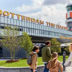 Rodzina zmierza na lotnisko Rotterdam The Hague Airport. W tle widoczna wieża kontroli lotów i napis z nazwą lotniska.
