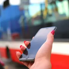 Dłoń z czerwonymi paznokciami trzyma smartfon, na którym można kupić bilet ZTM Warszawa online. W tle widać autobus.