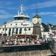 Tłum ludzi w kawiarni nad wodą w Oslo, z promem "Prinsen" i zegarem w tle. Ceny w Oslo mogą być wysokie, ale widok jest wart swojej ceny.