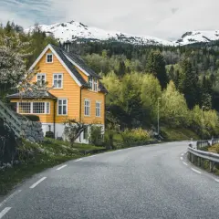Wjazd do Norwegii. Żółty dom z białymi oknami, kwitnące drzewo, droga wijąca się wśród zieleni i ośnieżone góry w tle.