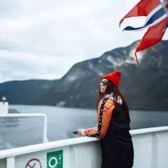 Kobieta na statku pod norweską flagą podziwia fiordy. Podróż po Norwegii, mimo że piękna, może wiązać się z wysokimi cenami.