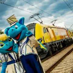 Dwa niebieskie smoki w strojach, obok znaku Energylandia, pozdrawiają przed pociągiem RMF. Idealny dojazd do Energylandii!