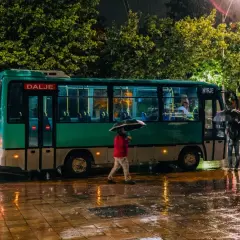 Deszczowa noc w Tirana Durres. Autobus z napisem "Dalje" i "Hyrje" stoi na mokrej ulicy, ludzie z parasolami czekają.