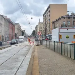 Ulica z torami tramwajowymi w remoncie, budynki mieszkalne, samochody i pracownicy w pomarańczowych kamizelkach.