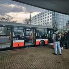 Praga autobus czeka na pasażerów przy nowoczesnym budynku. Grupa ludzi stoi na przystanku, gotowa do podróży.