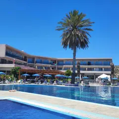 Basen i taras hotelu Cathrin Hotel Rodos z palmami i leżakami pod niebieskimi parasolami.