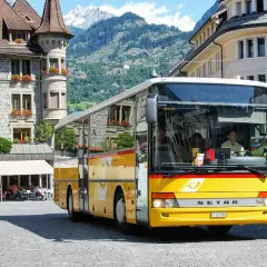 Żółty bus do Szwajcarii, z napisem "via Brigerbad", stoi na brukowanym placu przed kamiennymi budynkami i górami.
