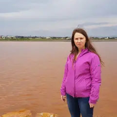 Kobieta w różowej kurtce stoi nad brzegiem Alicante różowego jeziora. W tle widać miasto.