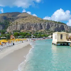 Słoneczna plaża w Palermo z turkusową wodą, górami w tle i budynkiem na palach.
