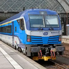Niebiesko-biały pociąg z żółtym pasem, gotowy do podróży do Czech, stoi na peronie pod nowoczesnym zadaszeniem dworca.