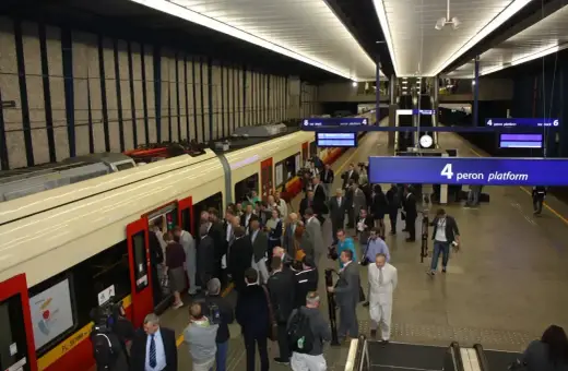 Ludzie wsiadają do pociągu na stacji Warszawa Centralna. Tablica informuje o peronie 4, który obsługuje połączenie do Lotniska Chopina.