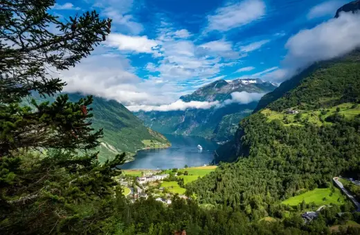 Majestatyczne fiordy Norwegii, zielone zbocza gór, błękitne niebo z chmurami i biały statek na wodzie.