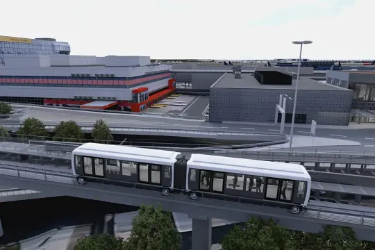Automatyczny pociąg na estakadzie na lotnisku Frankfurt nad Menem, z budynkami lotniska i samolotami w tle.