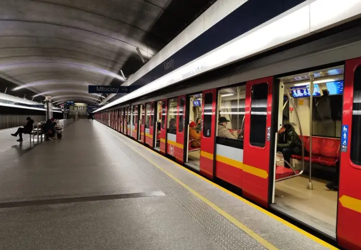 Stacja metra Młociny, do której jeździ metro w Warszawie. Czerwony pociąg z żółtym pasem stoi na peronie, czekając na pasażerów.