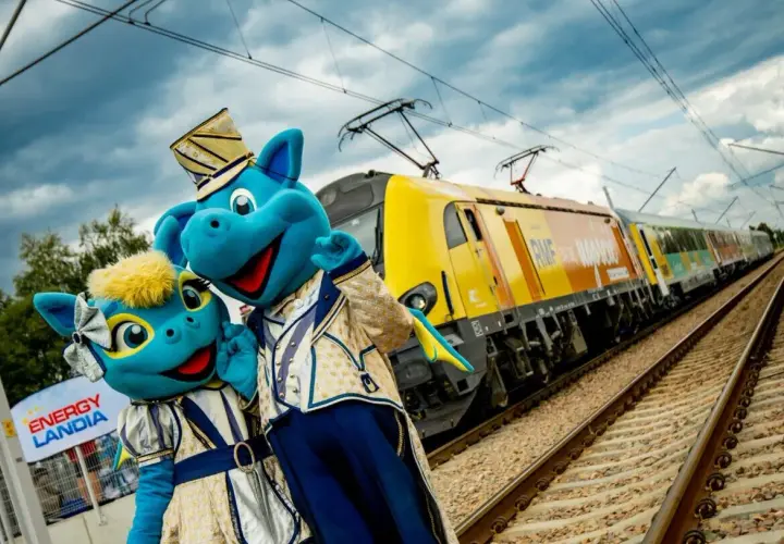 Dwa niebieskie smoki w strojach pozują przy żółtym pociągu. To pociąg do Energylandii, gotowy na podróż pełną wrażeń!