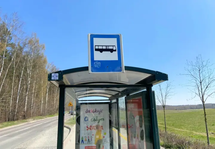 Szklane przystanki autobusowe z tabliczką informującą o transporcie publicznym, otoczone drzewami i zielonym polem.