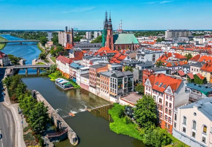 Widok na Opole z rzeką i grodem. Słoneczny dzień nad Odrą, widać kościół i kamienice.