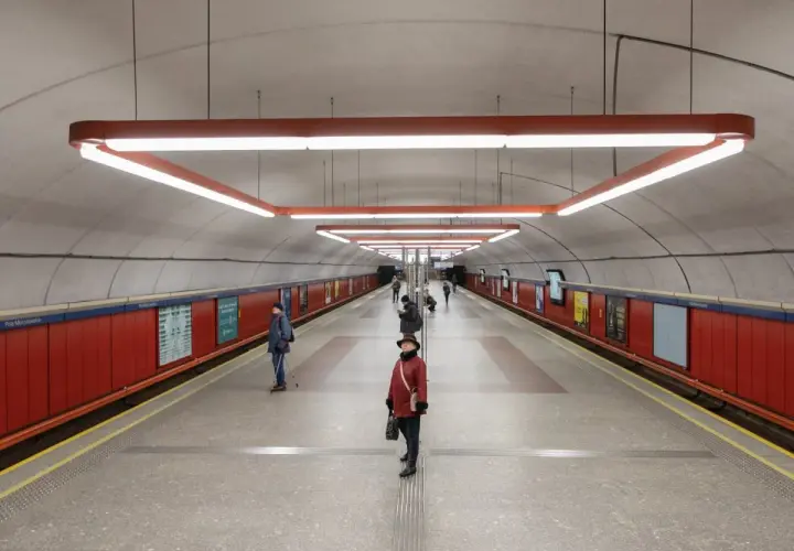 Stacja metra Pole Mokotowskie z nowoczesnym oświetleniem i czerwonymi panelami. Kilka osób czeka na peronie.