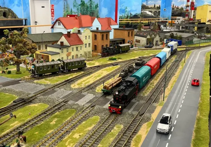 Miniaturowa kolejka z parowozem i wagonami, jak w Energylandii, przemierza malowniczy krajobraz z domkami i drogą.