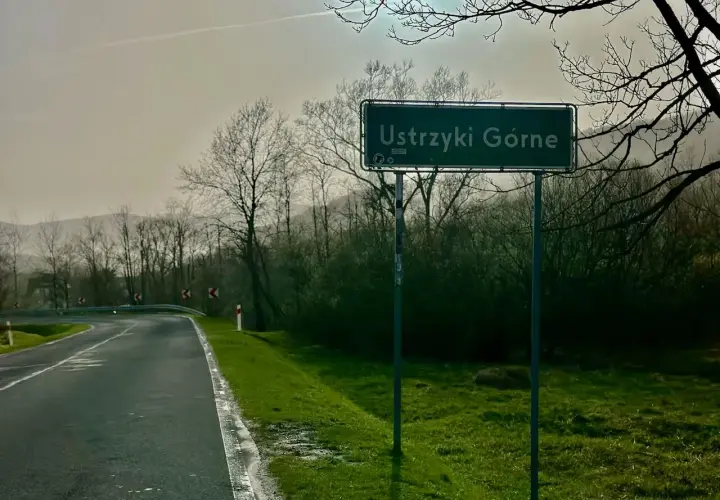 Droga z Krakowa do Ustrzyk Górnych. Znak z nazwą miejscowości Ustrzyki Górne na tle drzew i mglistego krajobrazu.