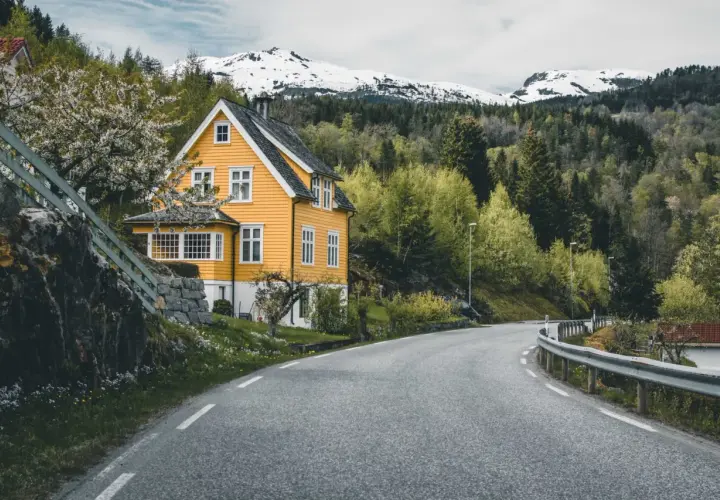 Wjazd do Norwegii. Żółty dom z białymi oknami, kwitnące drzewo, droga wijąca się wśród zieleni i ośnieżone góry w tle.