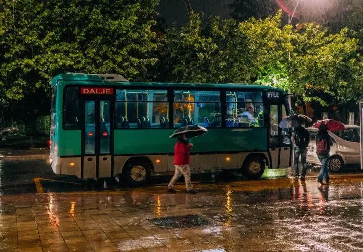 Deszczowa noc w Tirana Durres. Autobus z napisem "Dalje" i "Hyrje" stoi na mokrej ulicy, ludzie z parasolami czekają.