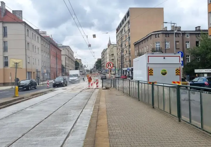 Ulica z torami tramwajowymi w remoncie, budynki mieszkalne, samochody i pracownicy w pomarańczowych kamizelkach.