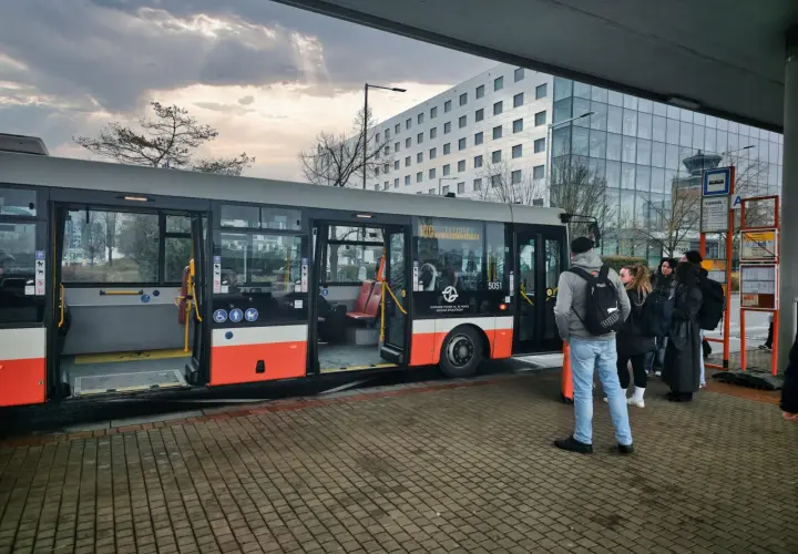 Praga autobus czeka na pasażerów przy nowoczesnym budynku. Grupa ludzi stoi na przystanku, gotowa do podróży.