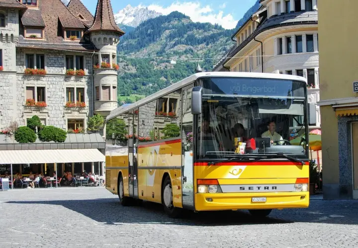 Żółty bus do Szwajcarii, z napisem "via Brigerbad", stoi na brukowanym placu przed kamiennymi budynkami i górami.