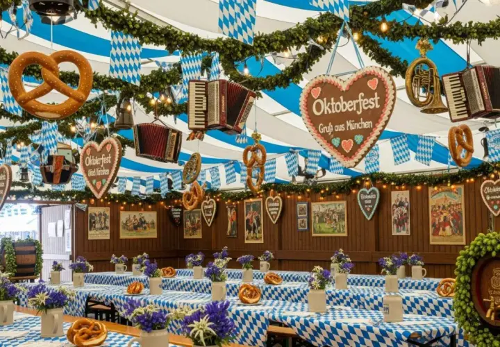 Wystrój namiotu na Oktoberfest w Monachium: wiszące precle, akordeony, serca z piernika i niebiesko-białe flagi. Stoły przykryte kratkowanymi obrusami, ozdobione kwiatami i beczką chmielu.