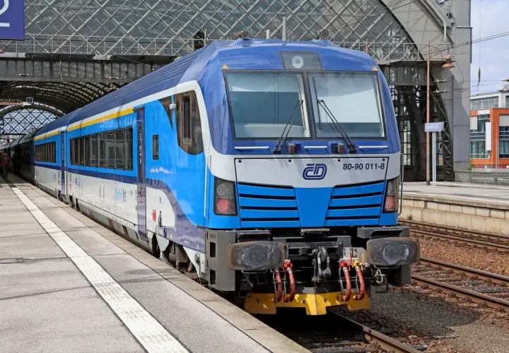 Niebiesko-biały pociąg z żółtym pasem, gotowy do podróży do Czech, stoi na peronie pod nowoczesnym zadaszeniem dworca.