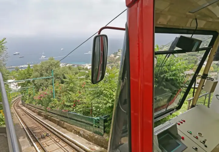 Czerwony wagonik kolejki linowej zjeżdża po stromym torze, oferując widok na zatokę pełną jachtów. To podróż jak z Neapolu do Sorrento, pełna zieleni i błękitu.