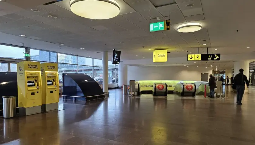 Dwa żółte automaty biletowe Arlanda Express, obok schodów ruchomych. Sprawdź arlanda express cena przed podróżą.