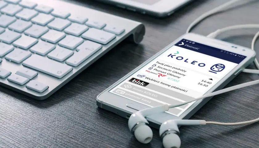 Smartfon z aplikacją KOLEO pokazuje plan podróży i opcje płatności, idealny do wyszukiwania zniżki PKP. Słuchawki i klawiatura sugerują planowanie podróży.