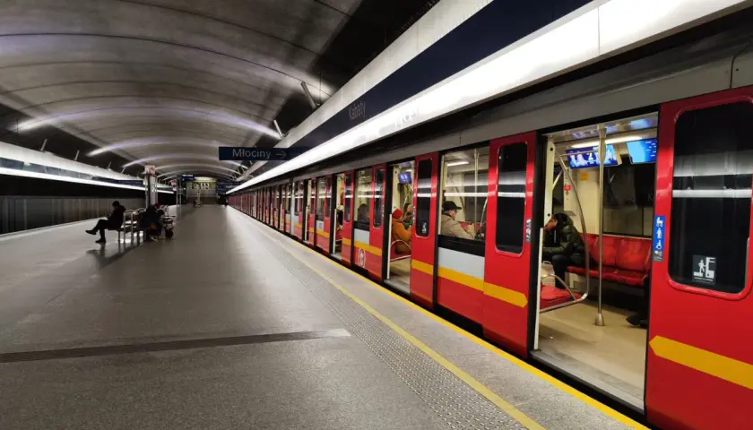 Stacja metra Młociny, do której jeździ metro w Warszawie. Czerwony pociąg z żółtym pasem stoi na peronie, czekając na pasażerów.