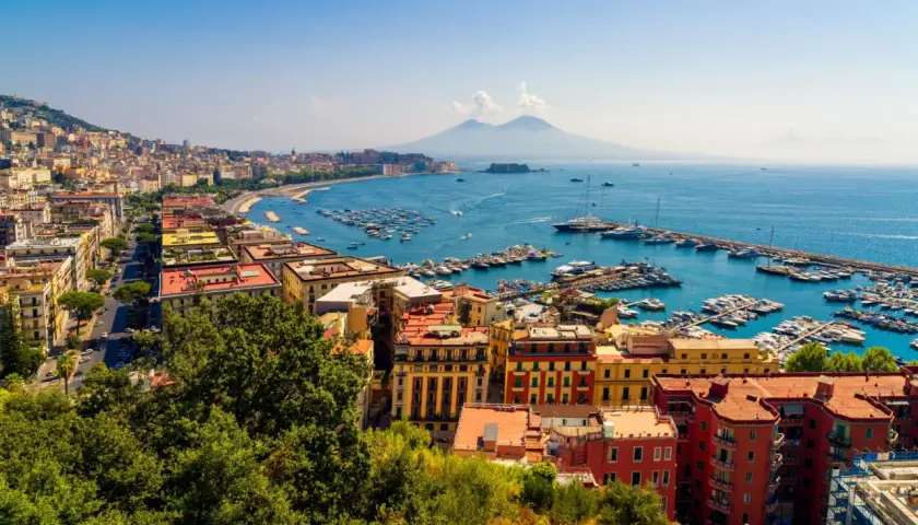 Widok na Zatokę Neapolitańską z Wezuwiuszem w tle. Miasto rozciąga się wzdłuż wybrzeża, a port pełen jest łodzi. Roma Napoli to marzenie wielu podróżników.
