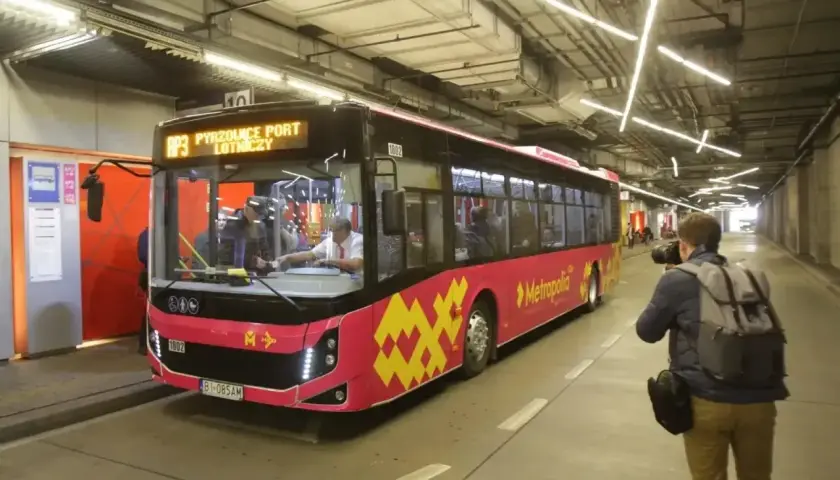 Różowy autobus linii AP3 z napisem "Pyrzowice Port Lotniczy" wjeżdża na pętlę. Obok stoi fotograf z aparatem. Bilety Katowice można kupić na pokładzie.