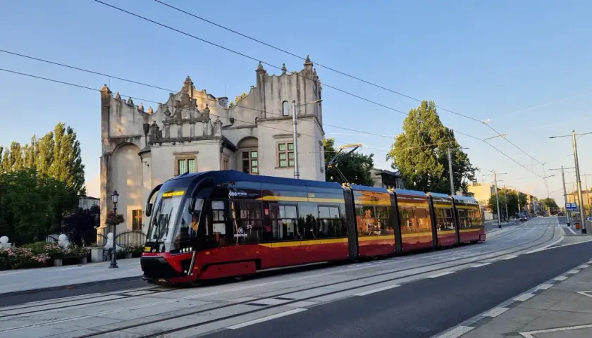 Nowoczesny tramwaj pabianice z czerwono-żółtymi pasami jedzie w stronę zabytkowego budynku.