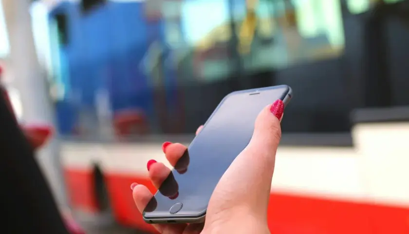 Dłoń z czerwonymi paznokciami trzyma smartfon, na którym można kupić bilety ZTM Warszawa. W tle widać autobus.