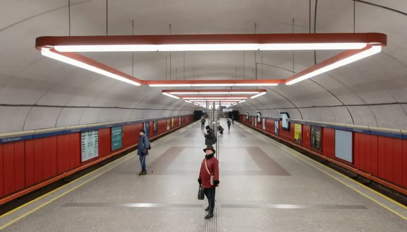Stacja metra Pole Mokotowskie z nowoczesnym oświetleniem i czerwonymi panelami. Kilka osób czeka na peronie.
