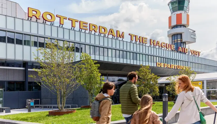Rodzina zmierza na lotnisko Rotterdam The Hague Airport. W tle widoczna wieża kontroli lotów i napis z nazwą lotniska.