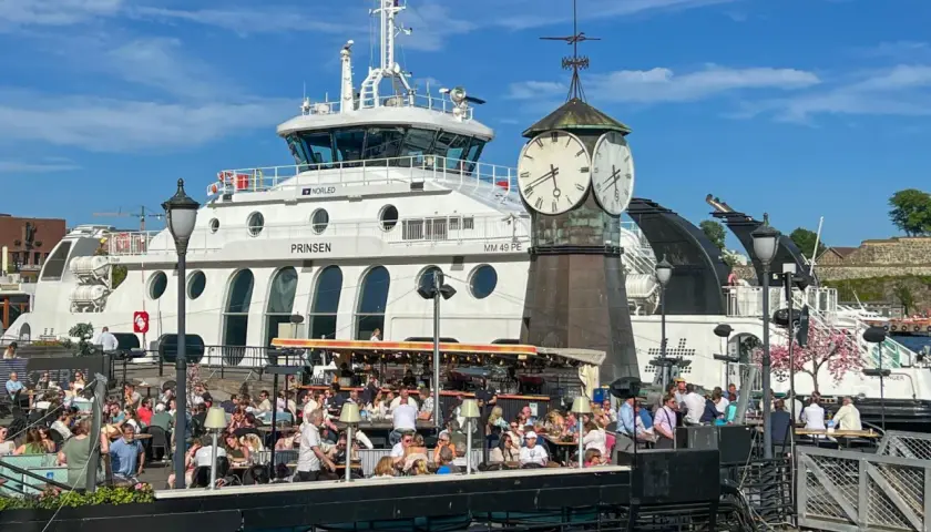 Tłum ludzi w kawiarni nad wodą w Oslo, z promem "Prinsen" i zegarem w tle. Ceny w Oslo mogą być wysokie, ale widok jest wart swojej ceny.